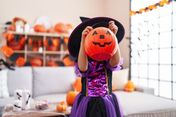 Adorable hispanic girl having halloween party holding pumpkin basket over face at home