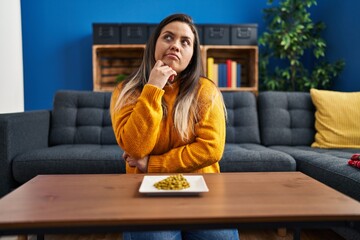 Young hispanic woman eating green peas thinking concentrated about doubt with finger on chin and...