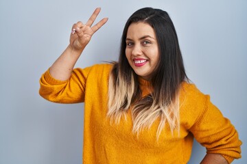 Young hispanic woman standing over isolated background smiling looking to the camera showing fingers doing victory sign. number two.