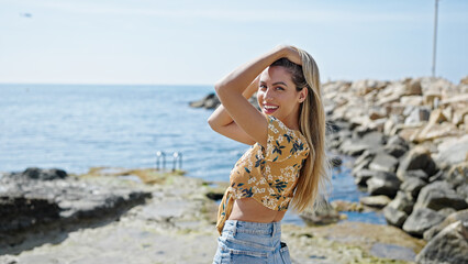 Young blonde woman smiling confident combing hair at seaside