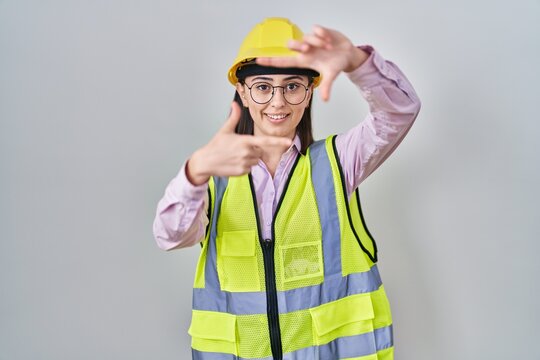 Hispanic girl wearing builder uniform and hardhat smiling making frame with hands and fingers with happy face. creativity and photography concept.
