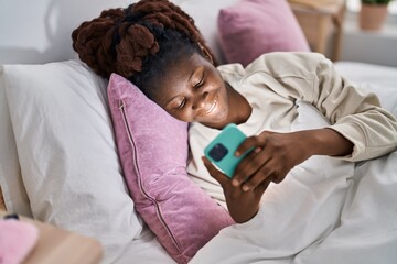 African american woman using smartphone lying on bed at bedroom