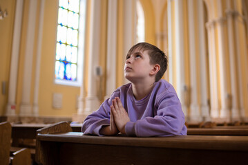a little boy in a jacket prays in a dark catholic church at a children's mass	