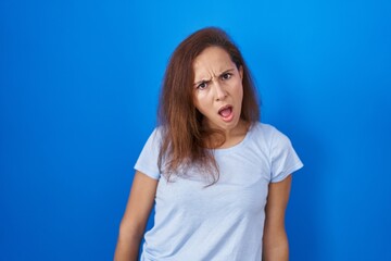 Brunette woman standing over blue background in shock face, looking skeptical and sarcastic, surprised with open mouth