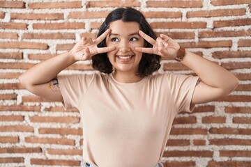 Young hispanic woman standing over bricks wall doing peace symbol with fingers over face, smiling cheerful showing victory