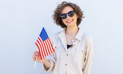 Young smiling happy fun woman in sunglasses hold US flag look camera on gray wall background. American travel.