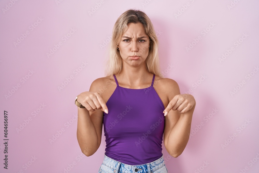 Poster Young blonde woman standing over pink background pointing down looking sad and upset, indicating direction with fingers, unhappy and depressed.