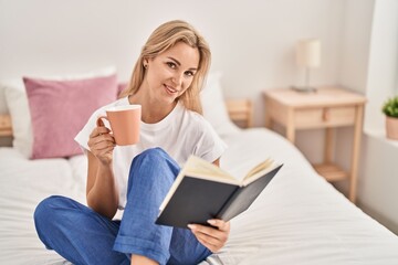 Young blonde woman drinking cup of coffee reading book at bedroom