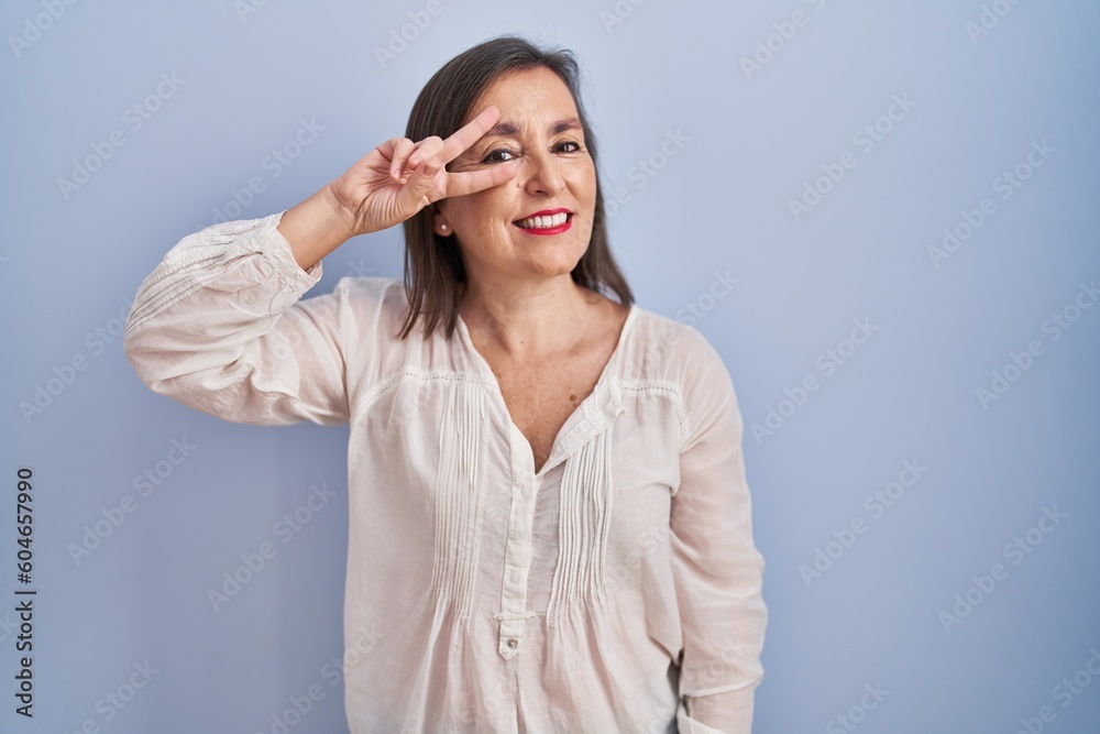 Canvas Prints middle age hispanic woman standing over blue background doing peace symbol with fingers over face, s