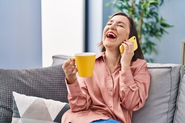 Fototapeta premium Middle age woman drinking coffee and talking on the smartphone at home
