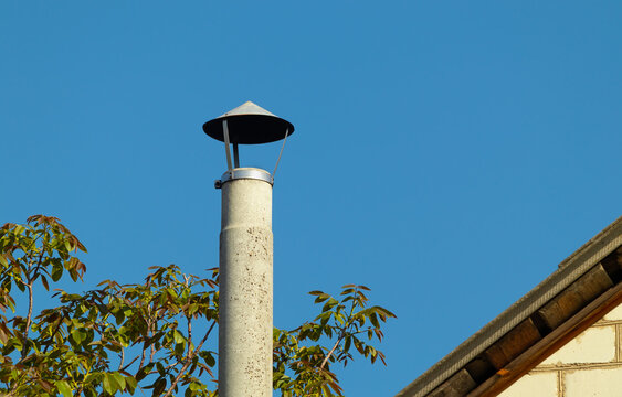 Concrete chimney pipe the edge of the roof of the house and the leaves on the branches against the background of a clear blue sky