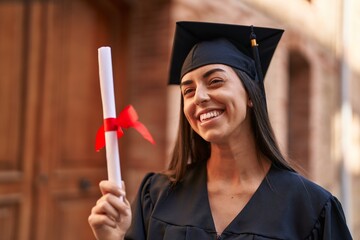 Young hispanic woman wearing graduated uniform holding diploma at university