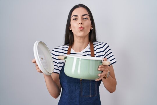 Young brunette woman wearing apron holding cooking pot looking at the camera blowing a kiss being lovely and sexy. love expression.