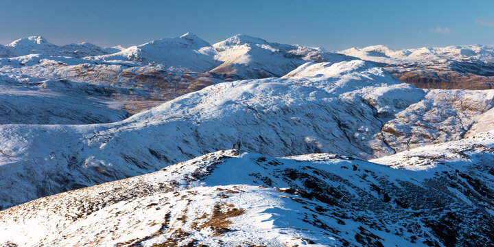 Two walkers hike over Ben Ledi in snow-capped winter mountain landscape, near Callander, Loch Lomond National Park, Scotland, UK
