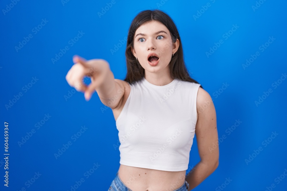 Sticker Young caucasian woman standing over blue background pointing with finger surprised ahead, open mouth amazed expression, something on the front