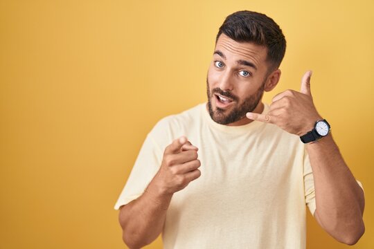Handsome hispanic man standing over yellow background smiling doing talking on the telephone gesture and pointing to you. call me.