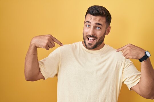 Handsome hispanic man standing over yellow background looking confident with smile on face, pointing oneself with fingers proud and happy.