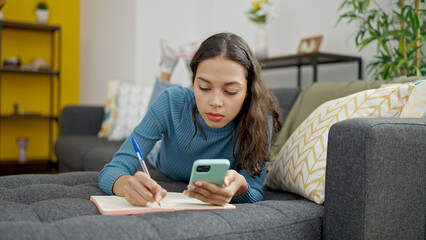 Young beautiful hispanic woman using smartphone writing on notebook lying on sofa at home