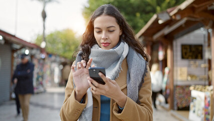 Young beautiful hispanic woman using smartphone smiling at street market