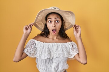 Young brazilian woman wearing summer hat over yellow background afraid and shocked with surprise and amazed expression, fear and excited face.