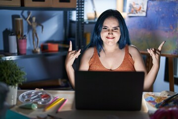 Young modern girl with blue hair sitting at art studio with laptop at night shouting with crazy expression doing rock symbol with hands up. music star. heavy concept.