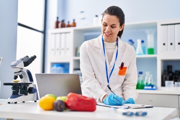 Young beautiful hispanic woman scientist using laptop writing on document at laboratory