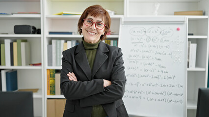 Mature hispanic woman teacher standing by white board with crossed arms at library university