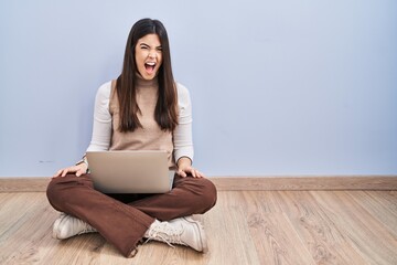 Young brunette woman working using computer laptop sitting on the floor angry and mad screaming frustrated and furious, shouting with anger. rage and aggressive concept.