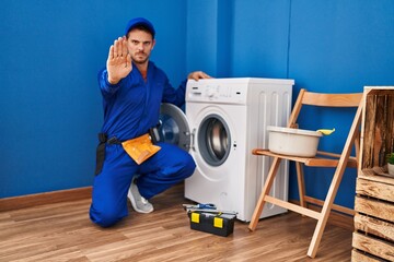 Young hispanic man working on washing machine with open hand doing stop sign with serious and confident expression, defense gesture