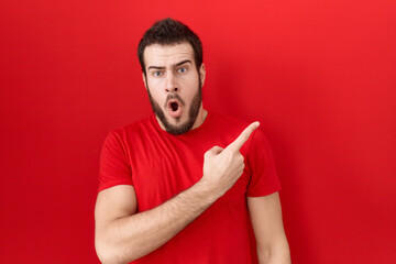 Young hispanic man wearing casual red t shirt surprised pointing with finger to the side, open mouth amazed expression.