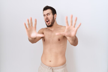 Young hispanic man standing shirtless over white background afraid and terrified with fear expression stop gesture with hands, shouting in shock. panic concept.