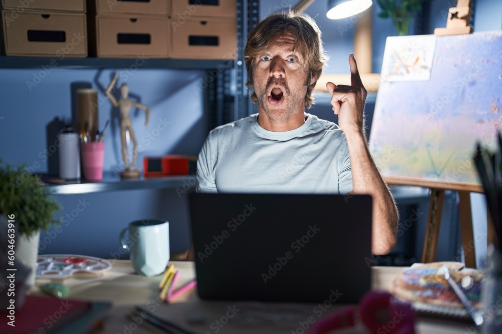 Sticker middle age man sitting at art studio with laptop at night pointing finger up with successful idea. e