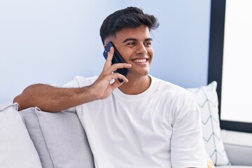 Young hispanic man talking on the smartphone sitting on sofa at home
