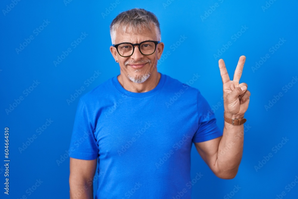 Wall mural Hispanic man with grey hair standing over blue background showing and pointing up with fingers number two while smiling confident and happy.