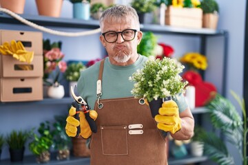 Hispanic man with grey hair working at florist shop puffing cheeks with funny face. mouth inflated...