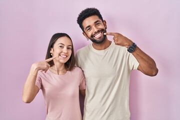 Young hispanic couple together over pink background smiling cheerful showing and pointing with fingers teeth and mouth. dental health concept.