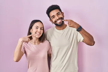 Young hispanic couple together over pink background smiling cheerful showing and pointing with fingers teeth and mouth. dental health concept.
