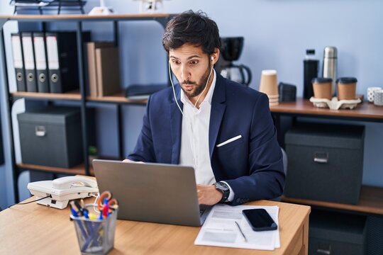 Handsome latin man working at the office using laptop scared and amazed with open mouth for surprise, disbelief face