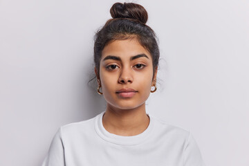 Portrait of beautiful serious brunette woman focused at camera has dark hair combed in bun dressed in casual t shirt isolated over white background. Confident Idian female model with calm expression - obrazy, fototapety, plakaty