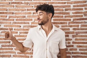 Arab man with beard standing over bricks wall background looking proud, smiling doing thumbs up gesture to the side