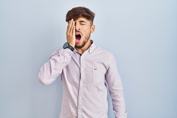 Arab man with beard standing over blue background yawning tired covering half face, eye and mouth with hand. face hurts in pain.