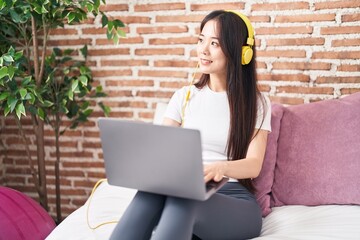 Young chinese woman using laptop and headphones sitting on bed at bedroom