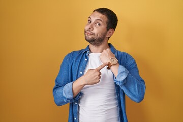 Hispanic man standing over yellow background in hurry pointing to watch time, impatience, looking...