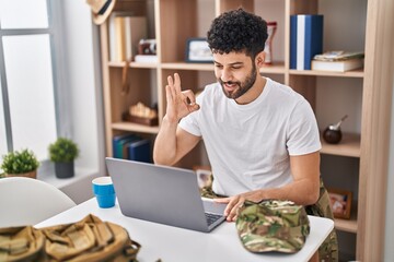 Arab man working as military doing video call with laptop doing ok sign with fingers, smiling friendly gesturing excellent symbol