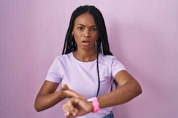 African american woman with braids standing over pink background in hurry pointing to watch time,...