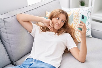 Young blonde girl using smartphone lying on sofa at home