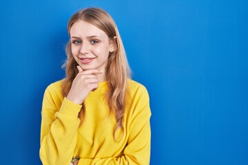 Young caucasian woman standing over blue background looking confident at the camera smiling with crossed arms and hand raised on chin. thinking positive.