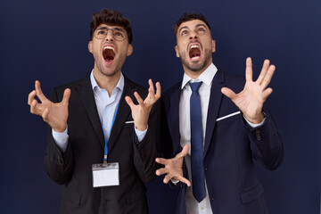 Two hispanic business men wearing business clothes crazy and mad shouting and yelling with aggressive expression and arms raised. frustration concept.