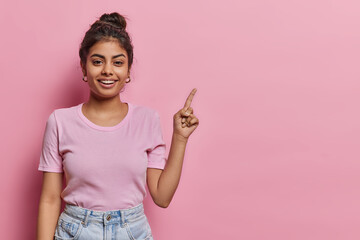 Attractive Indian teenage girl with dark hair points index finger above on copy space shows promotion offer demonstrates advertisement dressed in casual t shirt and jeans isolated over pink background - 604667975