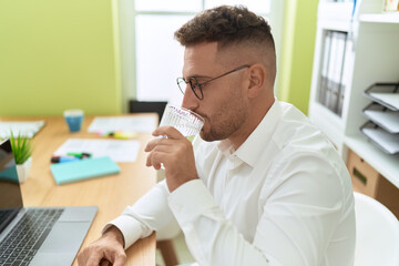 Young hispanic man business worker using laptop drinking water at office